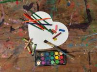 Art supplies on a wooden table, including colored markers, chalks, a paintbrush, and watercolor paints, arranged around a white paper heart.