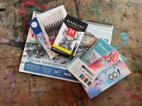 Various art supplies including watercolor paper, oil pastels, sketch pencils, and a drawing pad.